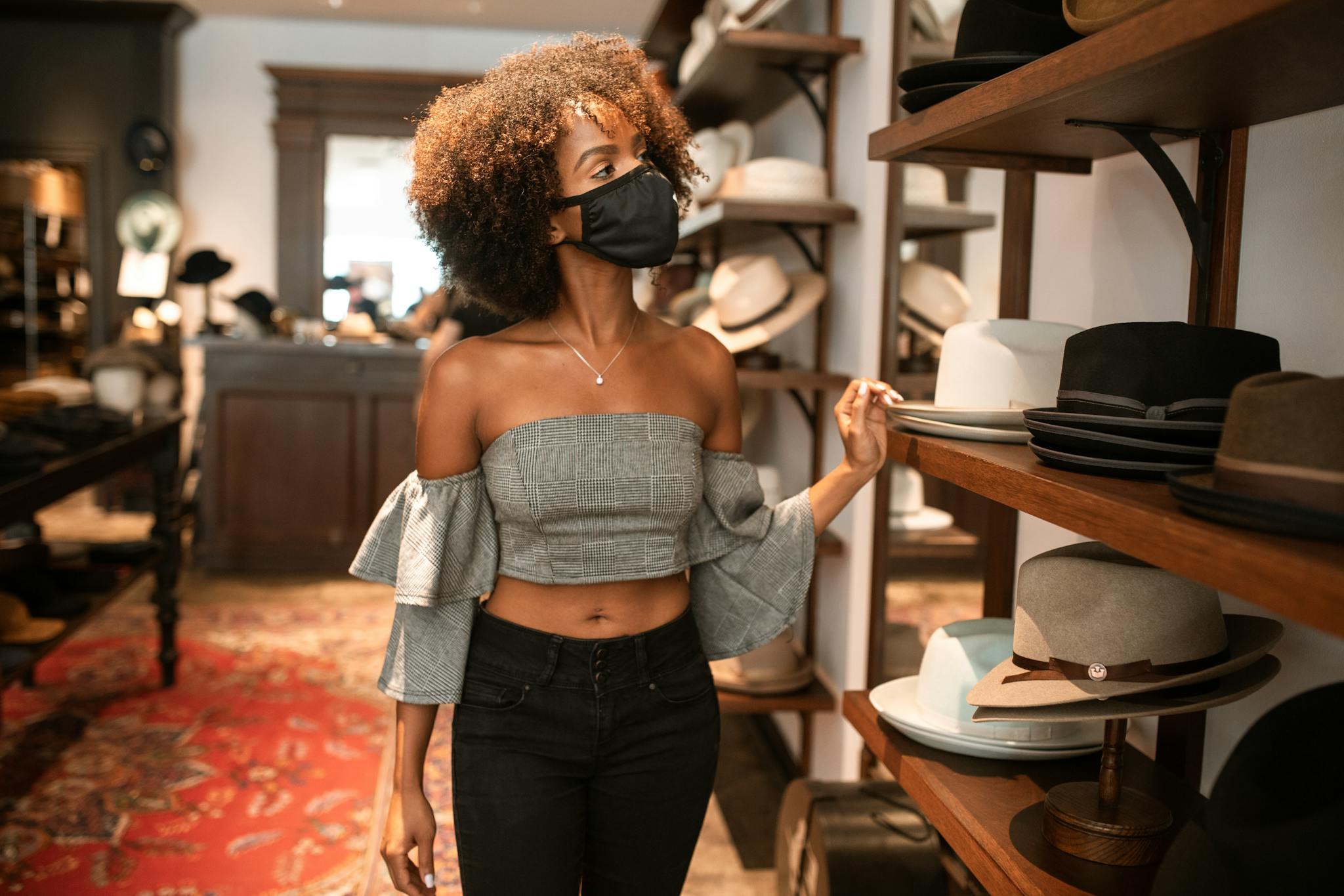 Elegant woman with curly hair and face mask browsing hats in chic store.